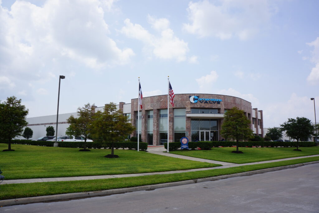 Cornerstone Valve office building with a modern design, featuring two flagpoles displaying the American and Texas flags, surrounded by a neatly maintained green lawn and trees under a partly cloudy sky.